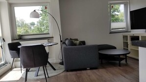 Moderni olohuone, jossa on harmaa sohva, pyöreä pöytä, lattiavalaisin ja televisio, joka on sijoitettu lipaston päälle.