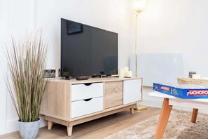 A modern living room with a flat-screen TV, a wooden cabinet, a potted plant, a standing lamp, and a Monopoly board game. - City Escape: Chic 1 Bed Apt in Central Leeds (Leeds)