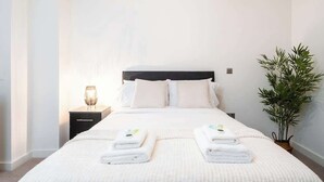 Una cama bien hecha con dos toallas dobladas, una mesita de noche con una lámpara y una planta en maceta.