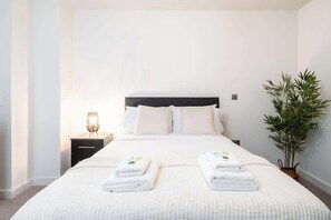 A neatly made bed with two folded towels, a bedside table with a lamp, and a potted plant.
