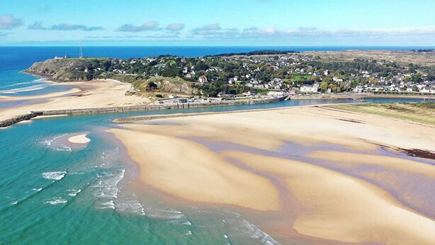 An aerial view of a coastal area with a sandy beach, a small town in the background, and a lighthouse on a hill.