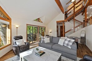 Una sala de estar moderna con un sofá gris, un sillón de cuero y una mesa de centro de mármol. Se observa una escalera de madera con barandillas de vidrio, y un gran ventanal que da a un espacio exterior.