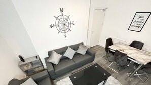 Un soggiorno moderno con un divano grigio, cuscini decorati, un tavolino da caffè nero, un tappeto bianco, un tavolo da pranzo con sedie e una decorazione murale a forma di bussola.