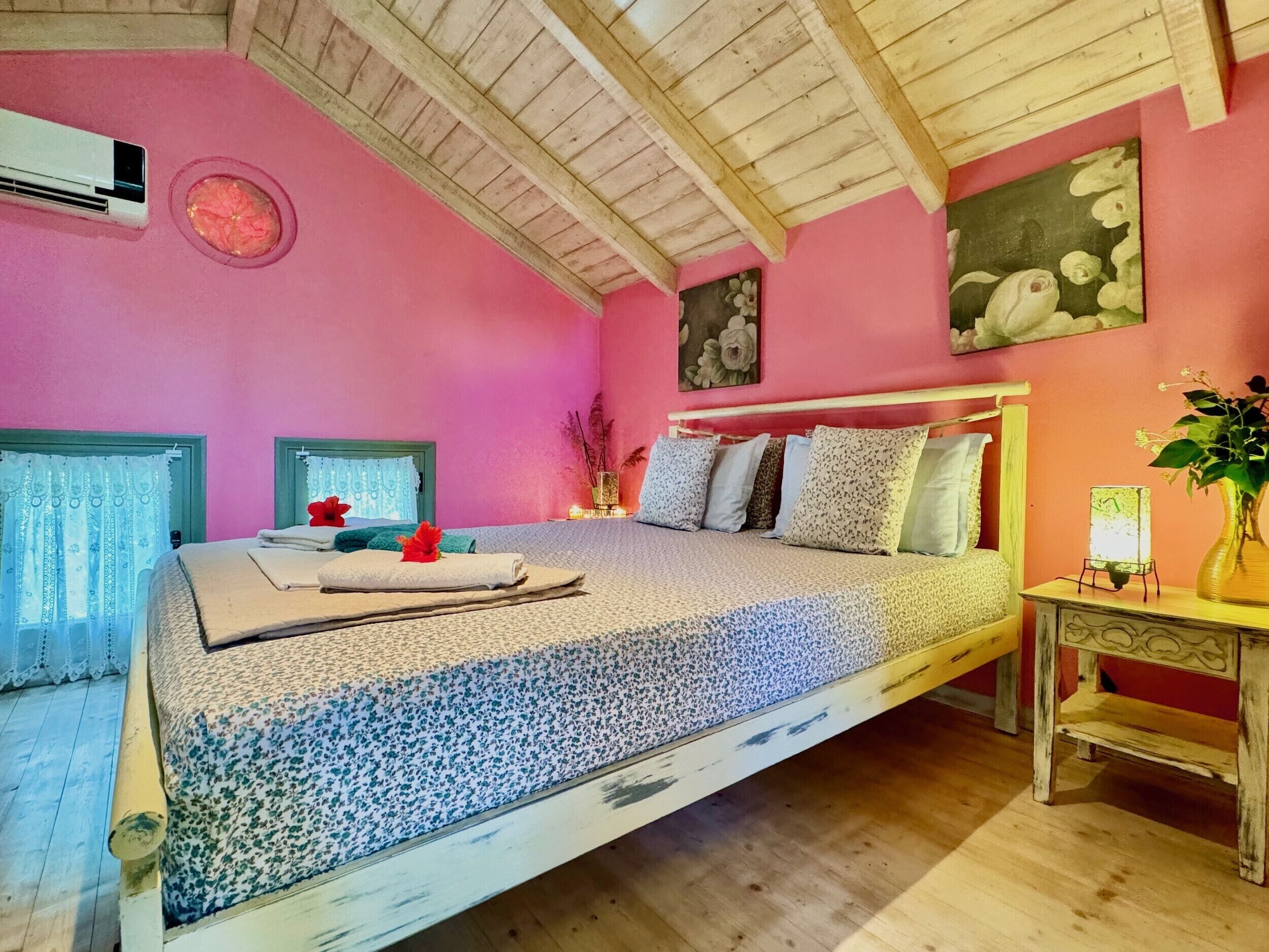 Une chambre avec un plafond en bois, des murs roses, un lit avec une literie à motifs floraux et une table de chevet sur laquelle se trouve un vase.
