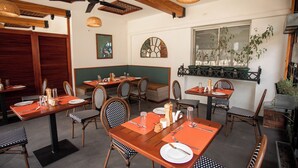A restaurant with wooden tables and chairs, a ceiling fan, and a large window with plants. - The Choate Boutique Hotel & Restaurant (Kampala)