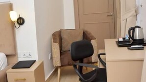 Habitación de hotel pequeña con cama, escritorio con silla, mesita de noche y lámpara de pared, además de una puerta.