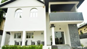 Una casa moderna de dos pisos con un balcón, un patio cubierto con mobiliario exterior y una escalera que conduce a la entrada.