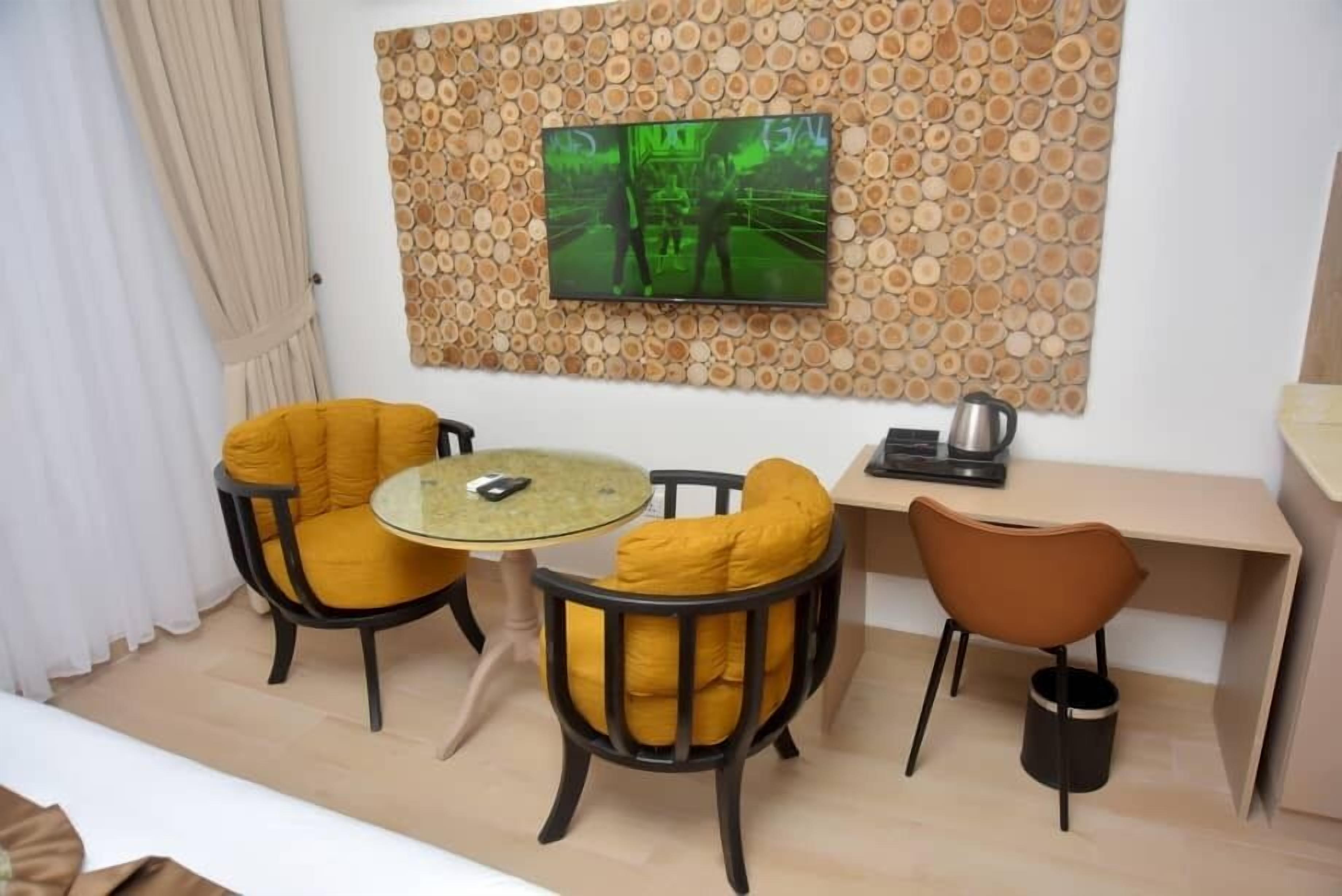 A modern room with a round glass table, two yellow cushioned chairs, a brown chair, a desk with a kettle, and a TV mounted on a wood-patterned wall.
