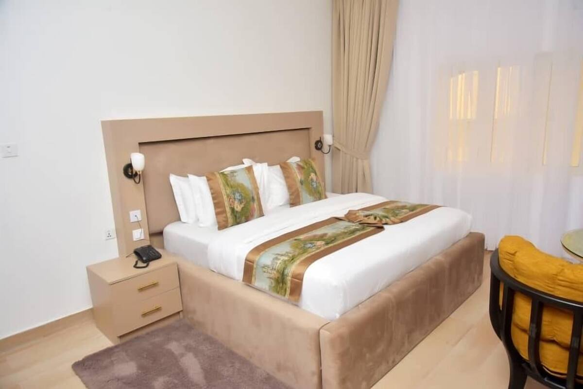 Ein Schlafzimmer mit einem großen Bett, einem Nachttisch mit Telefon, einer kleinen Schublade, einem braunen Teppich und einem Stuhl mit gelbem Kissen.