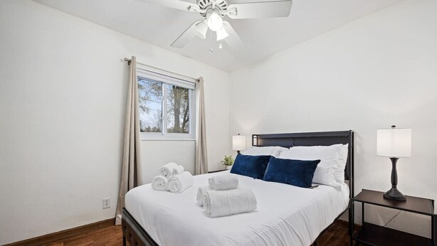 A bedroom with a bed, a nightstand, a ceiling fan, a window with curtains, and a view of trees outside. - Poolside Palisade - A Fun Family Retreat for 6 (Marquette Heights)