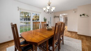 A dining room with a wooden table and chairs, a chandelier, and a view through large windows. - Poolside Palisade - A Fun Family Retreat for 6 (Marquette Heights)