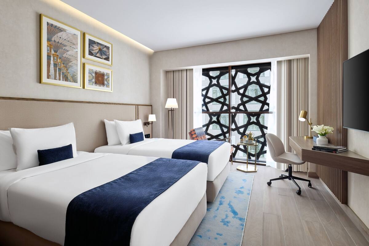 A hotel room with two beds, a desk, a chair, a TV, and a decorative wall panel.