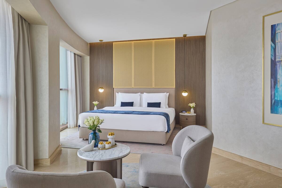 A modern hotel room with a large bed, two bedside tables, a round coffee table, and two armchairs.