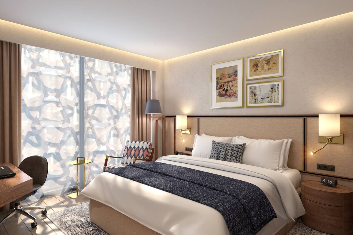 A modern hotel room with a large bed, a desk, and a chair. There are two wall-mounted lamps and a window with patterned curtains.