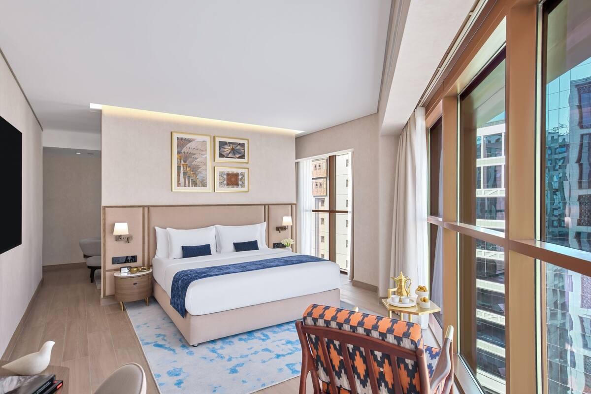 A hotel room with a large bed, bedside tables, a small table with a lamp, a chair, and a view of the city through large windows.