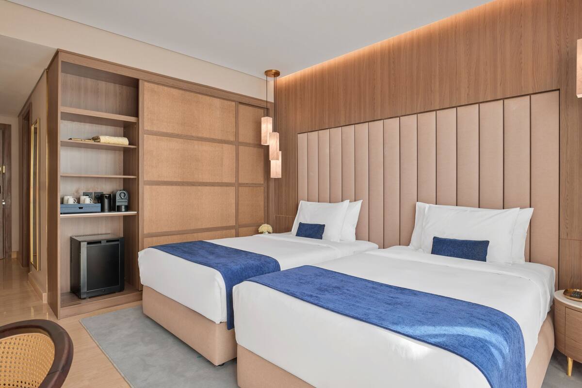 A hotel room with two beds, a minibar, a desk, and a large window.