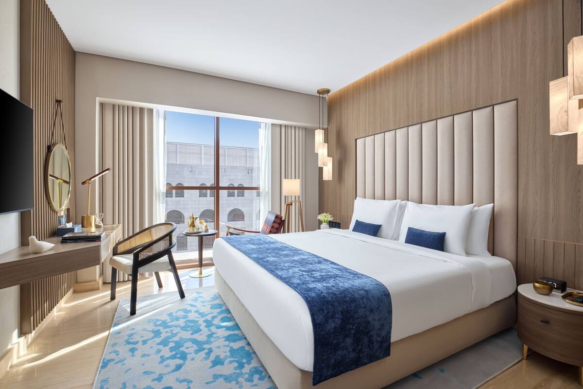A modern hotel room with a large bed, a desk, a chair, and a view of the city.