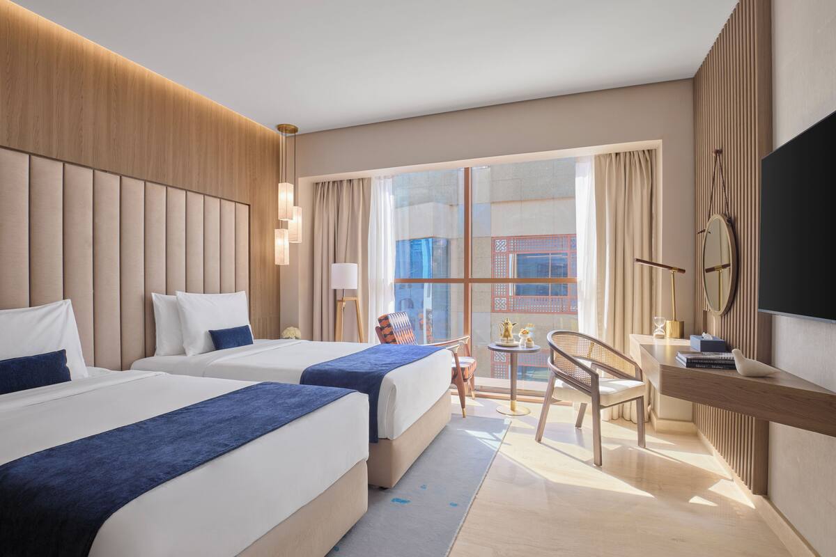 A hotel room with two beds, a desk, a chair, a TV, and a view of the city.