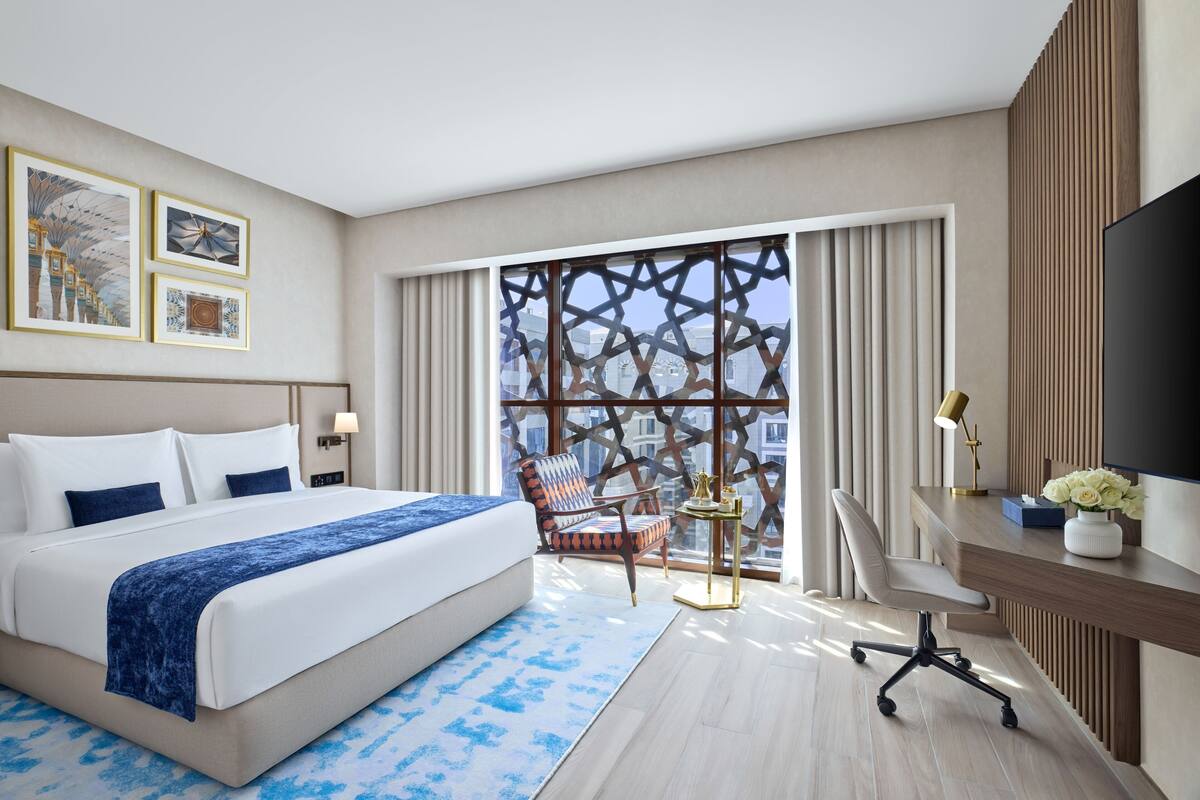 A hotel room with a large bed, a desk with a TV, a chair, and a window with a view of buildings.