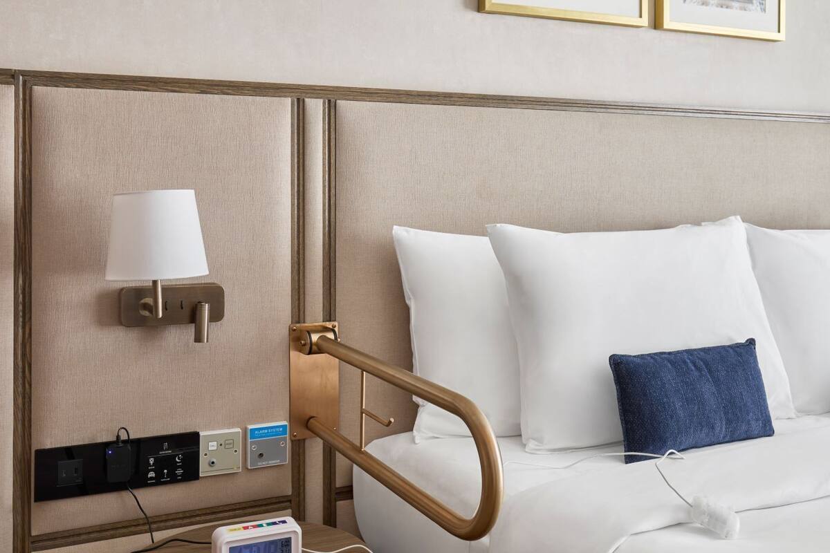 A hotel room with a bed, bedside table, lamp, clock, and artwork on the wall.