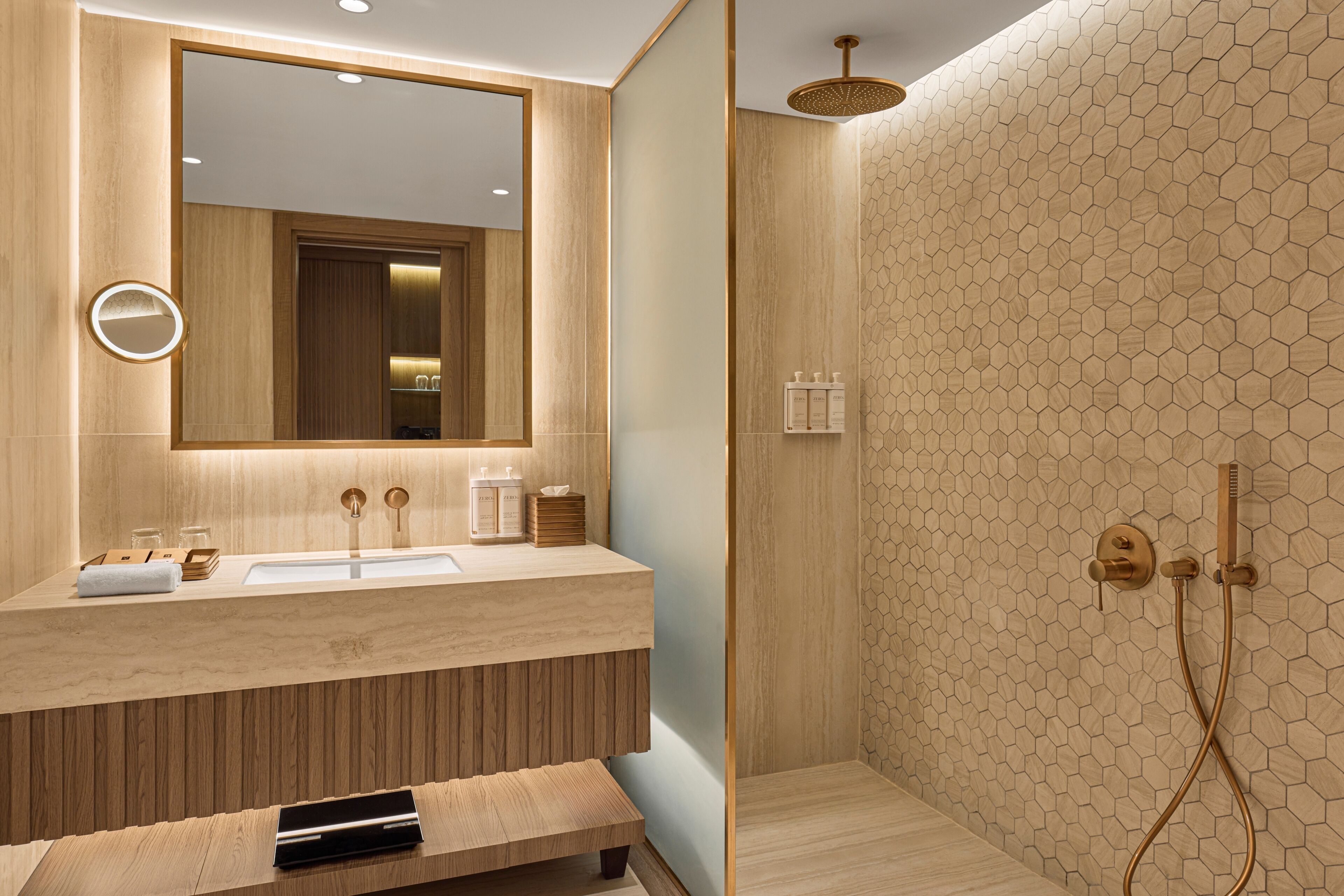 A modern bathroom with a wooden vanity, a large mirror, and a shower with a hexagonal tile wall.