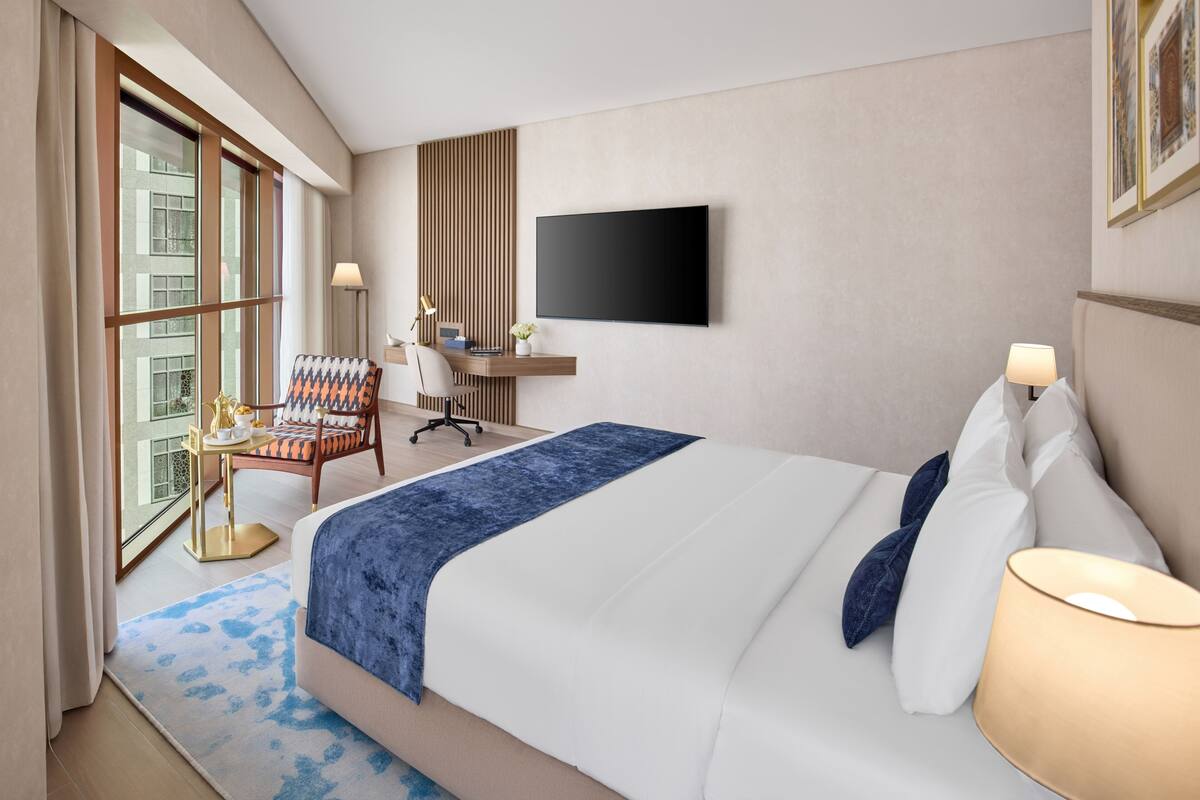 A hotel room with a large bed, a desk with a chair, a TV mounted on the wall, and a window with curtains.