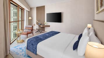 A hotel room with a large bed, a desk with a chair, a TV mounted on the wall, and a window with curtains.
