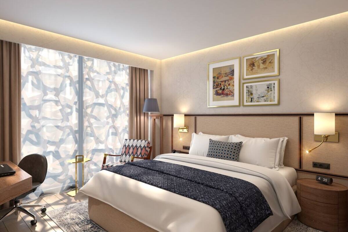 A modern hotel room with a large bed, a desk, and a chair. There are two wall-mounted lamps and a window with curtains.