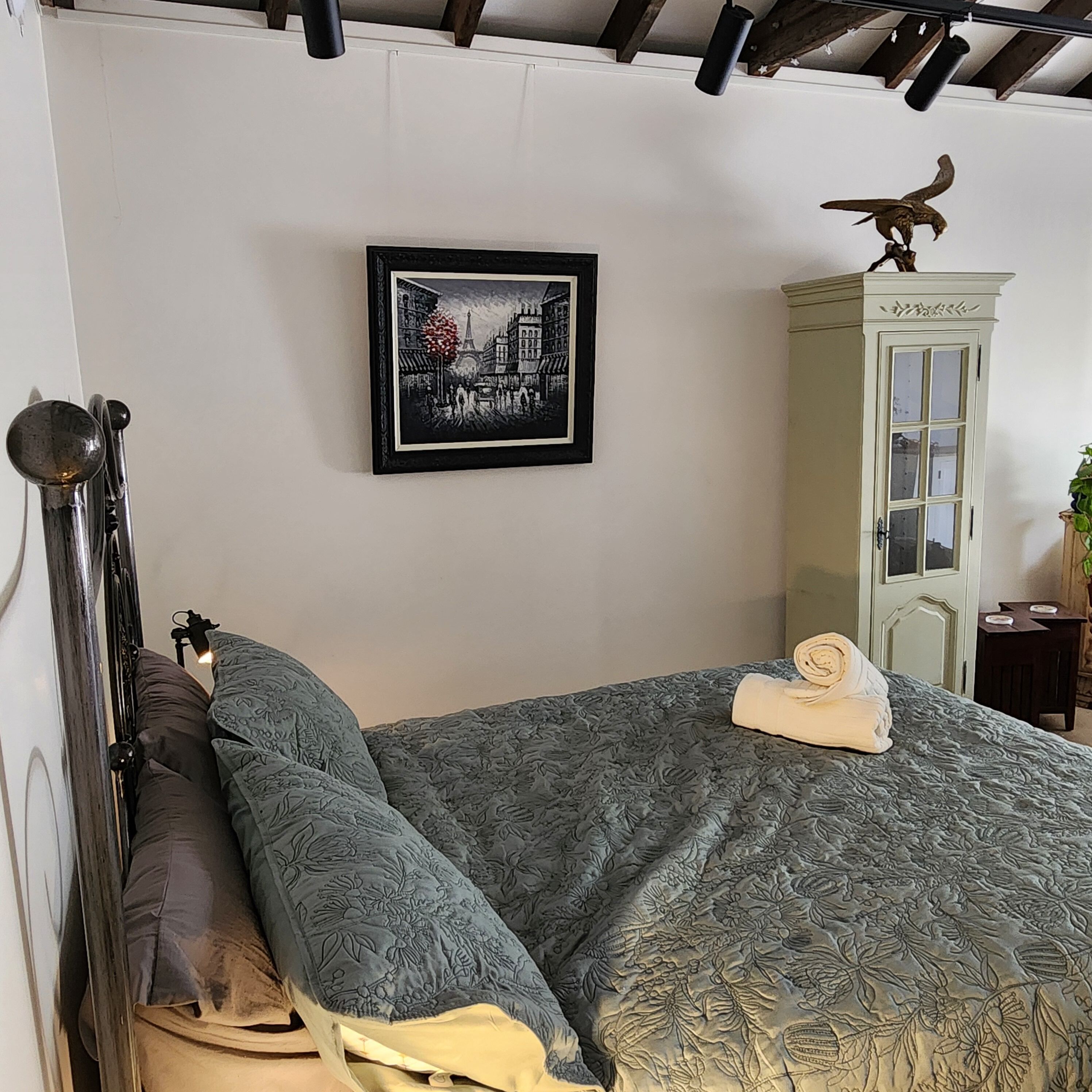 A neatly made bed with a patterned bedspread, a framed picture of a cityscape on the wall, and a cabinet with a bird sculpture on top.