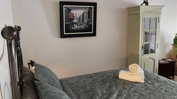 A neatly made bed with a patterned bedspread, a framed picture of a cityscape on the wall, and a cabinet with a bird sculpture on top.