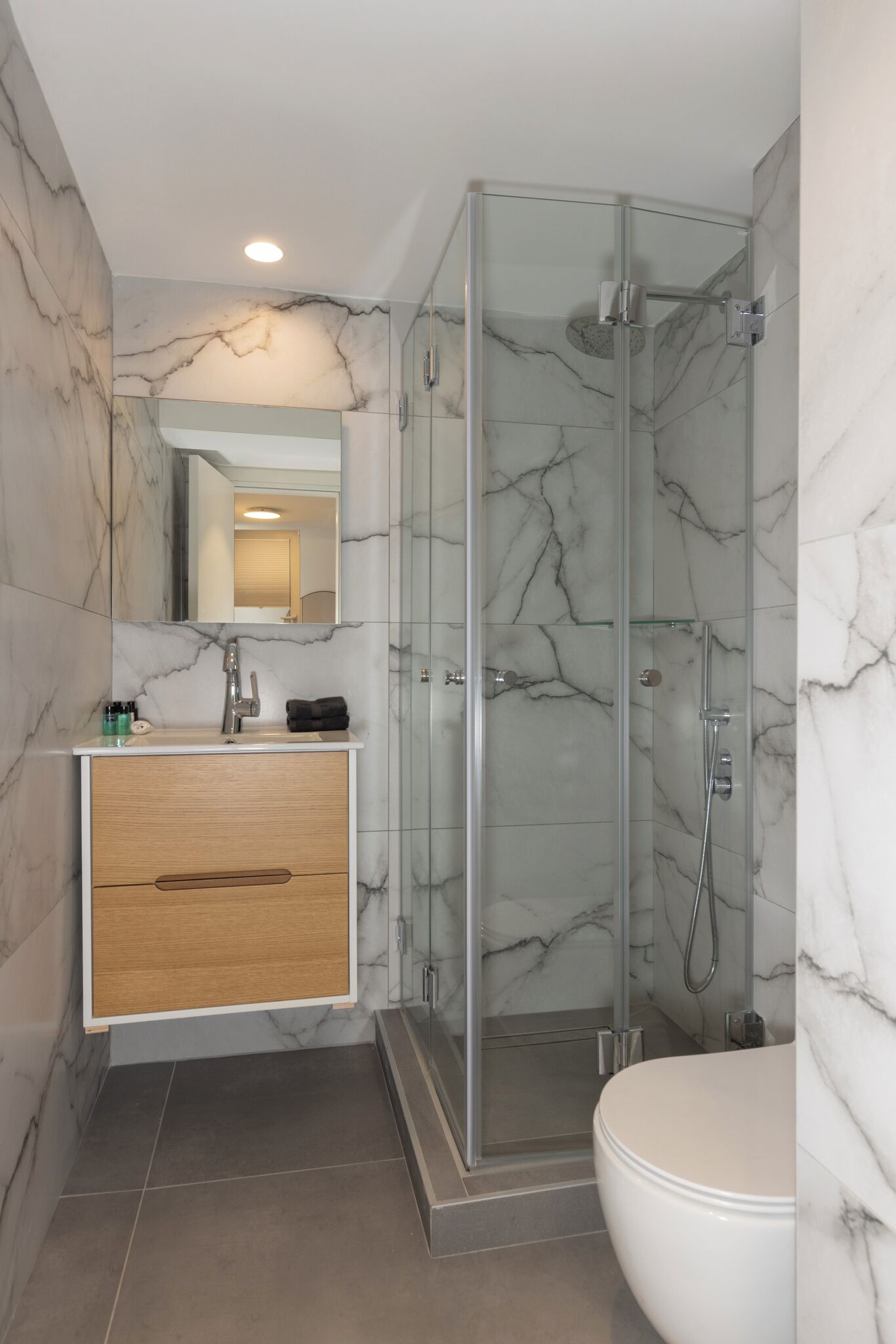 Een moderne badkamer met een marmeren wand, een douchecabine van glas, een houten wastafelmeubel en een wit toilet.