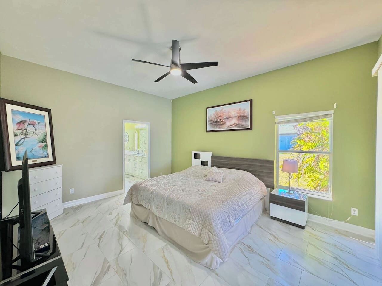 A bedroom with a bed, a ceiling fan, a TV, a window with a view, and a nightstand.