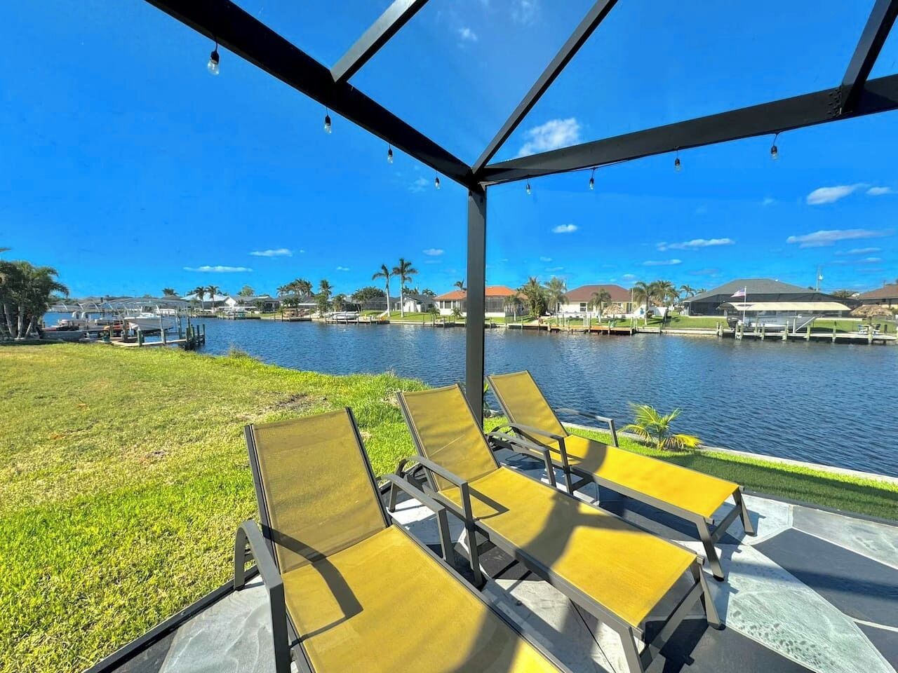 A covered outdoor seating area with chairs facing a body of water, surrounded by a grassy area and buildings in the background.