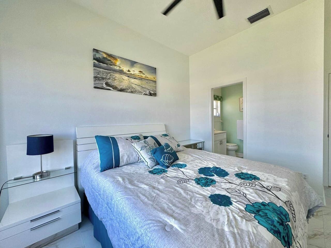 Un dormitorio con cama, mesitas de noche, lámpara, un cuadro en la pared y un baño visible a través de una puerta abierta.