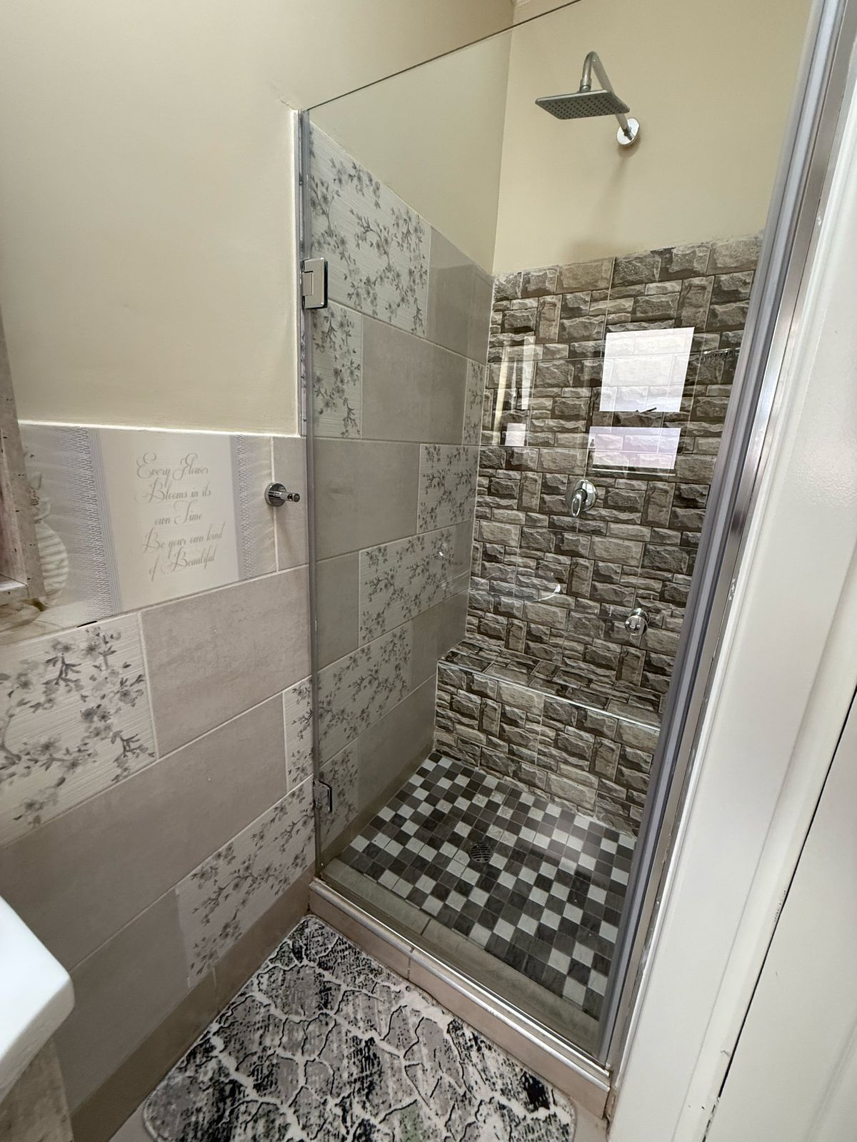 A shower with a glass door, stone wall, and checkered floor tiles.