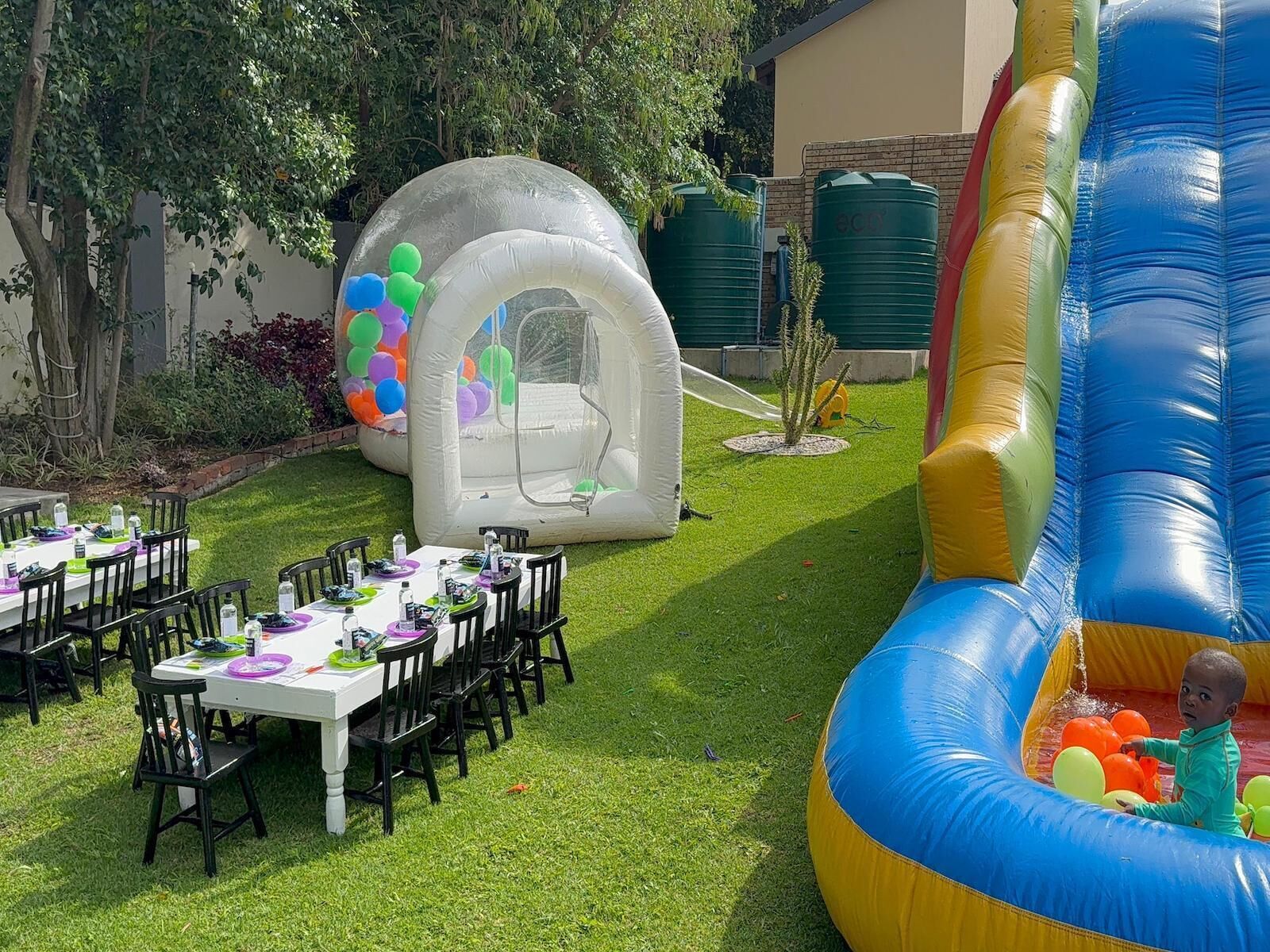 Un espai a l'aire lliure preparat per a una festa, amb una taula de dinar, cadires i jocs inflables.