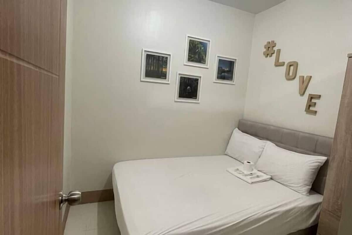 A bedroom with a bed, a wall-mounted shelf with framed photos, a #LOVE sign, and a door.