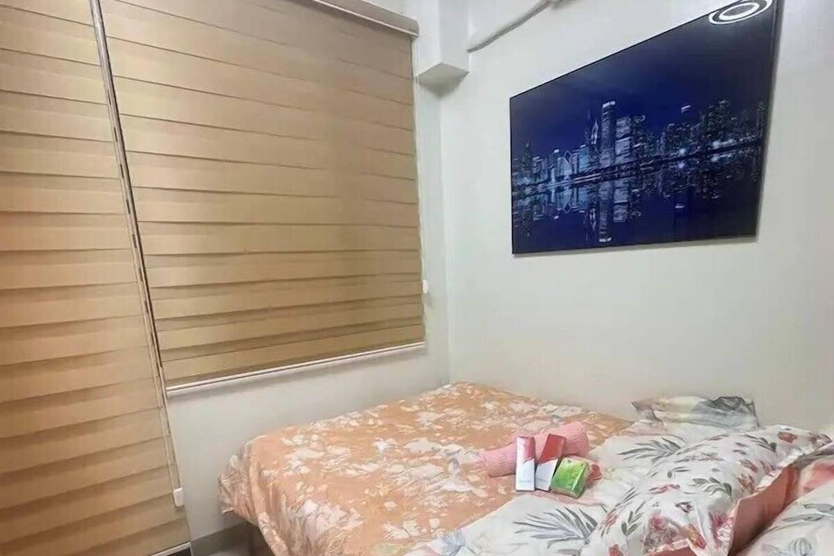 A bedroom with a bed, a nightstand, a window with blinds, a wall-mounted air conditioner, and a cityscape painting.