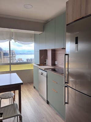 Uma cozinha moderna com máquina de lavar louça embutida, frigorífico de aço inoxidável e vista para a paisagem urbana através de grandes janelas.