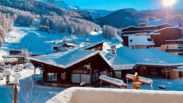 A snow-covered mountain resort with wooden buildings, a ski lift, and a clear view of the surrounding mountains.