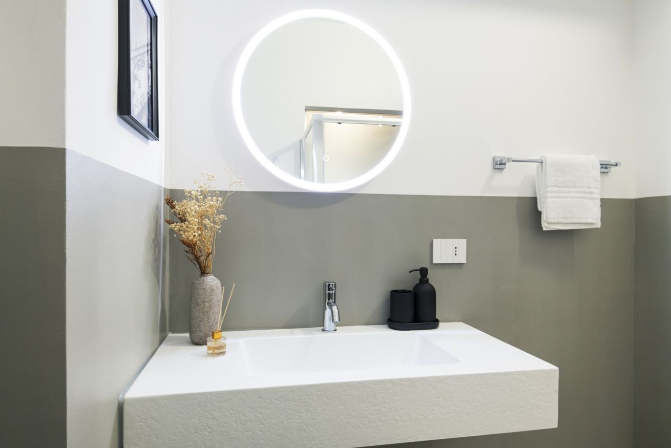 Un baño moderno con un lavamanos blanco, un espejo redondo con luz, un florero con flores secas y un perchero para toallas.