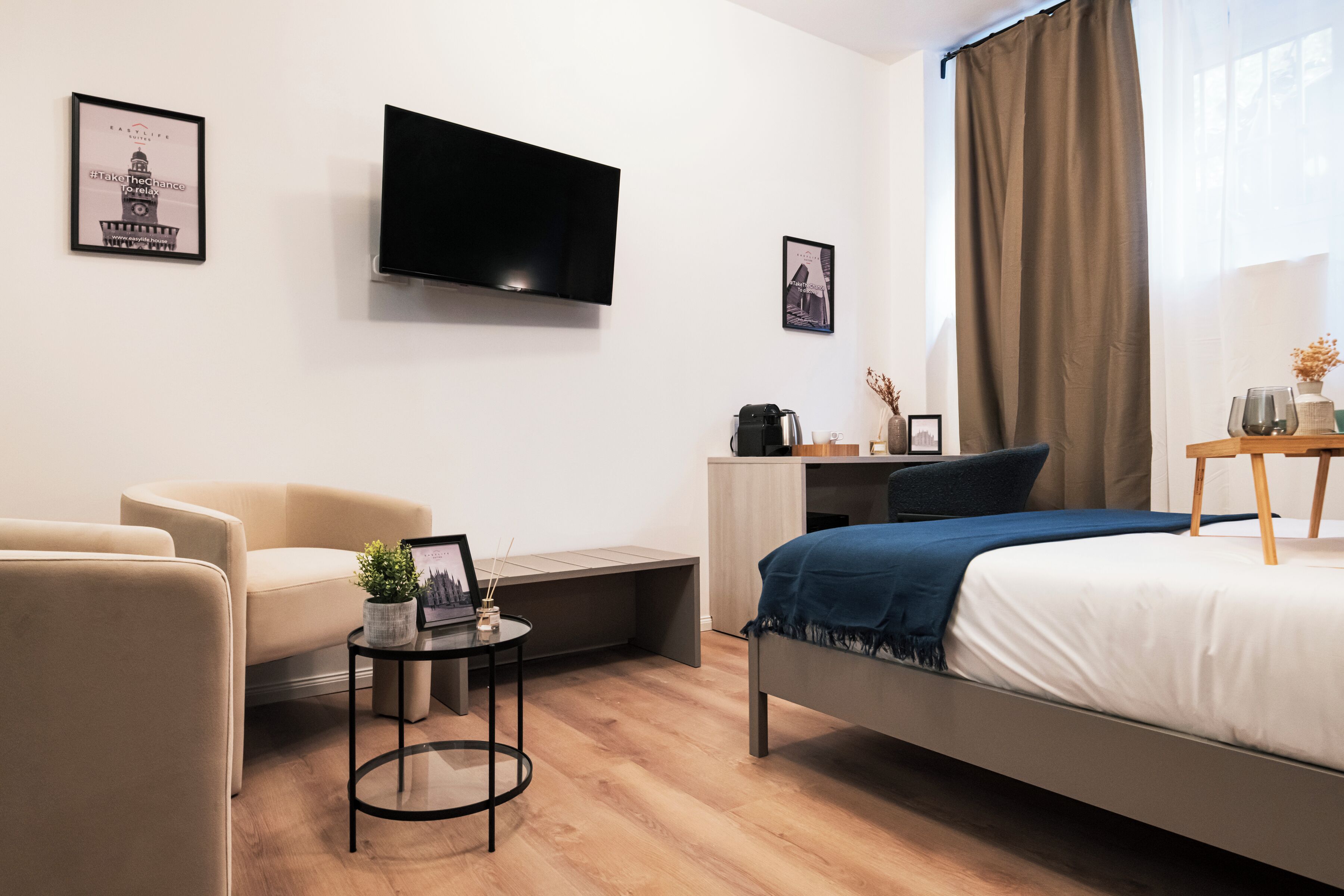 A modern hotel room with a bed, a desk, a chair, a TV, and a small table with a plant.