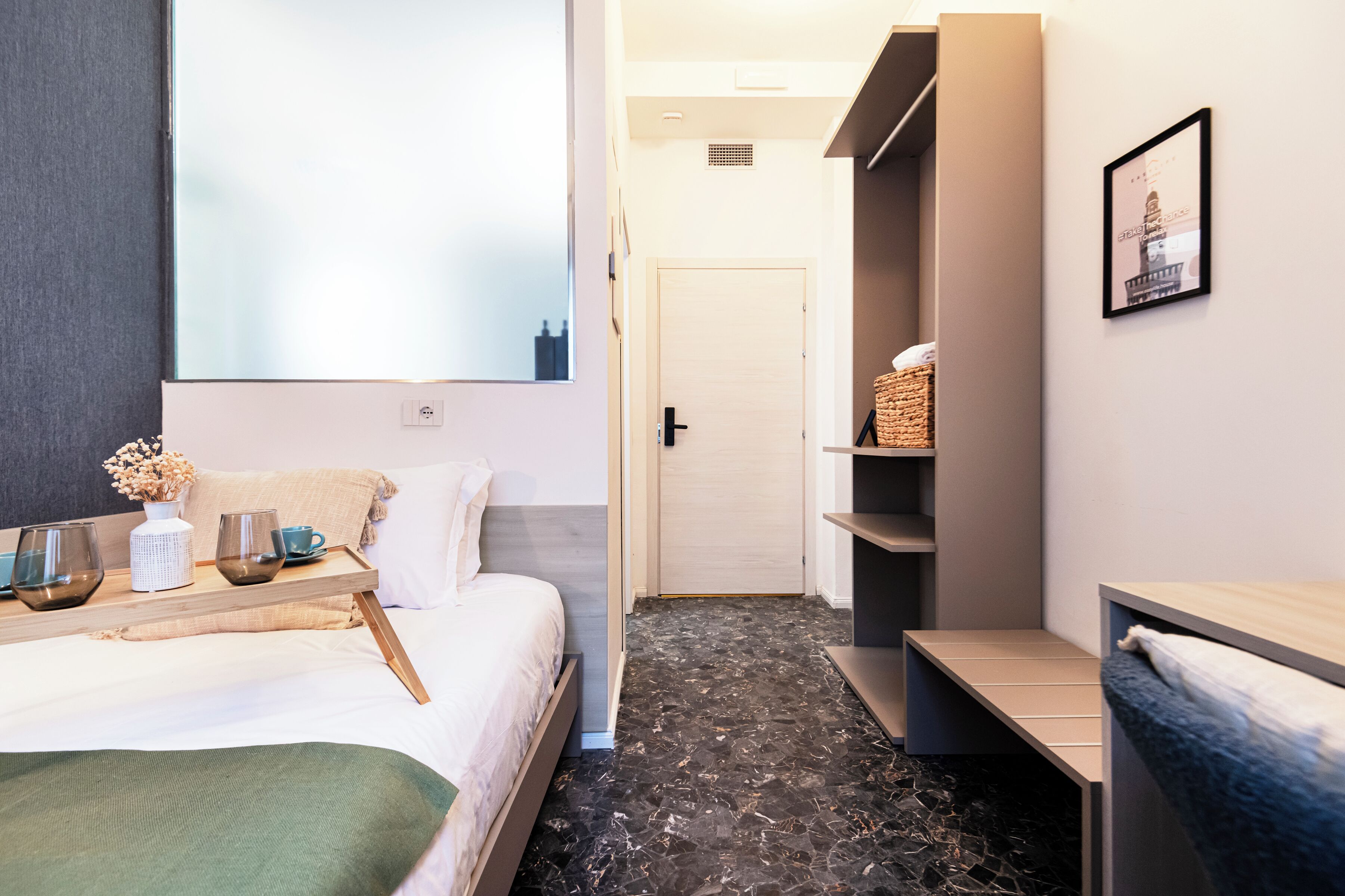 A modern hotel room with a bed, bedside table, and a large window. There is a door leading to another room and a shelf with items on the wall.