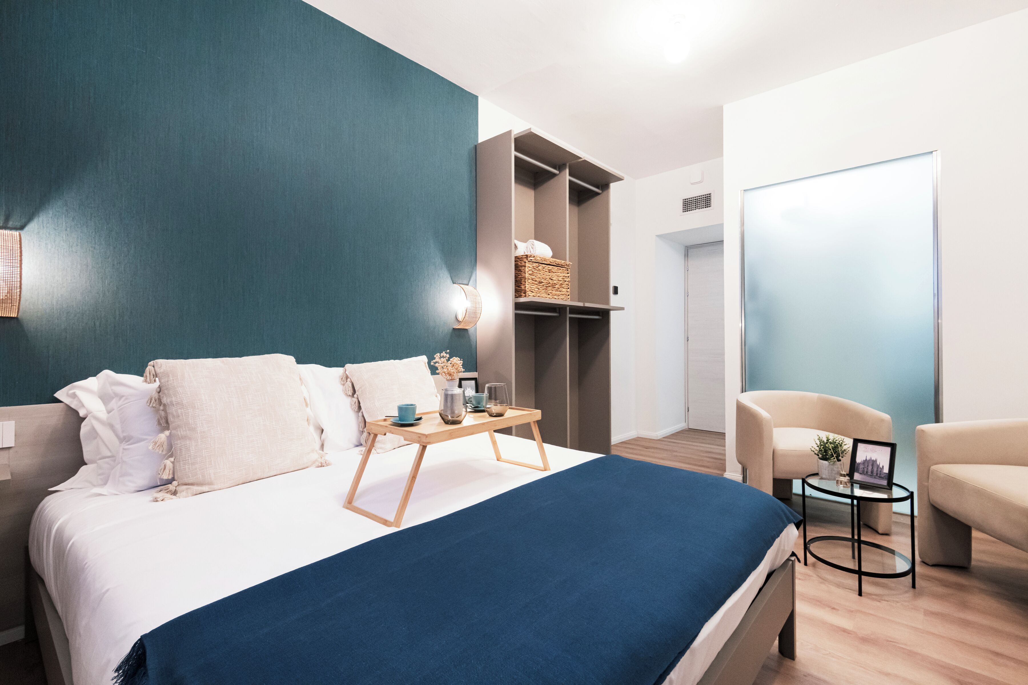 A modern hotel room with a bed, bedside tables, a seating area with chairs, and a wardrobe.