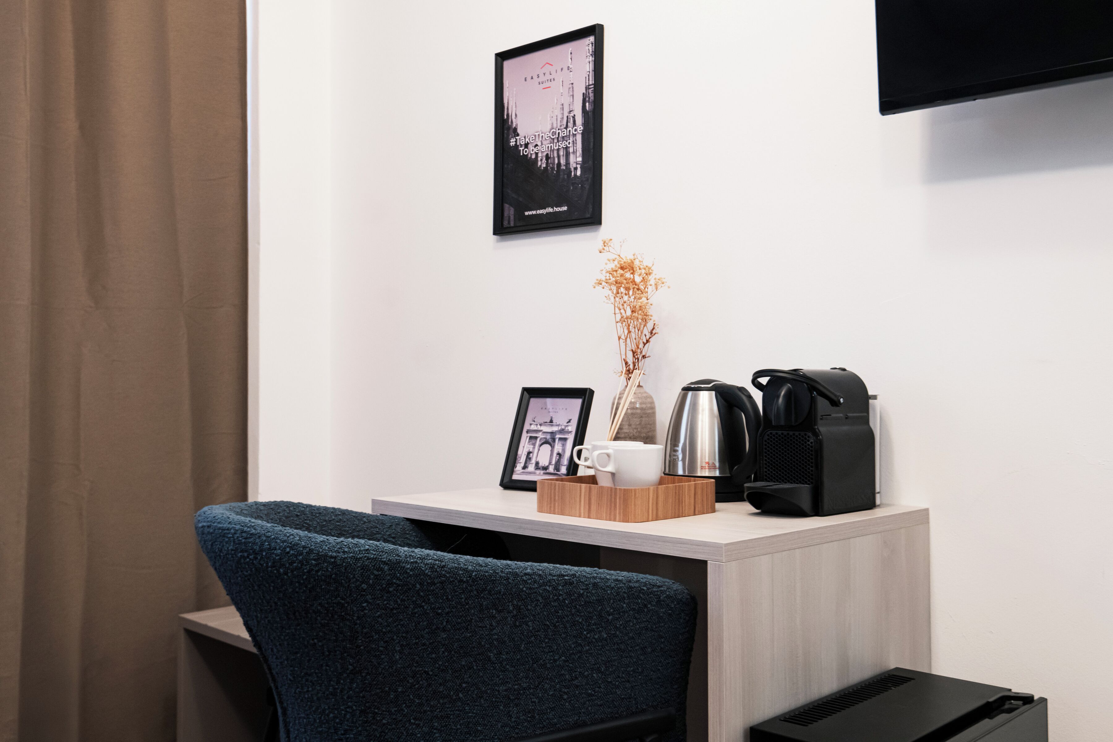 A hotel room with a desk, a framed picture, a coffee maker, and a vase with dried flowers.