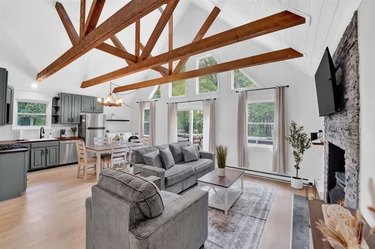 Ein großzügiger Wohnbereich mit hoher Decke, freiliegenden Holzbalken, einem grauen Sofa, einem Esstisch und einem Kamin.