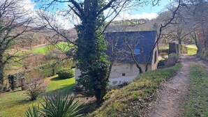 A rustic stone house with a sloped roof, surrounded by a rural landscape with a dirt path leading to it.