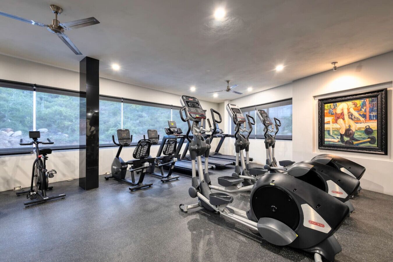 A gym with various exercise machines, large windows, and a ceiling fan.