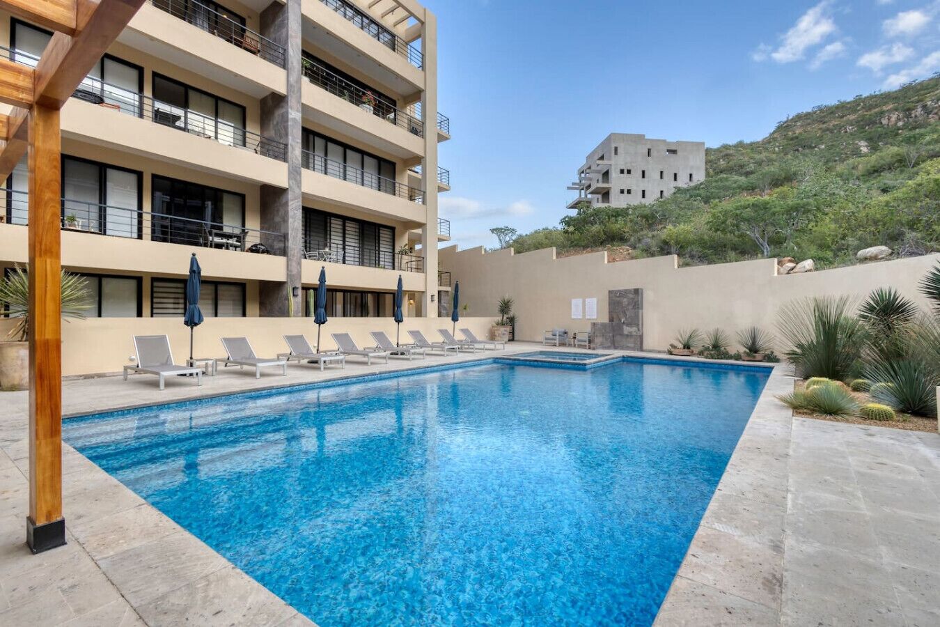 A swimming pool with lounge chairs and umbrellas, surrounded by a building with balconies and a hillside in the background.