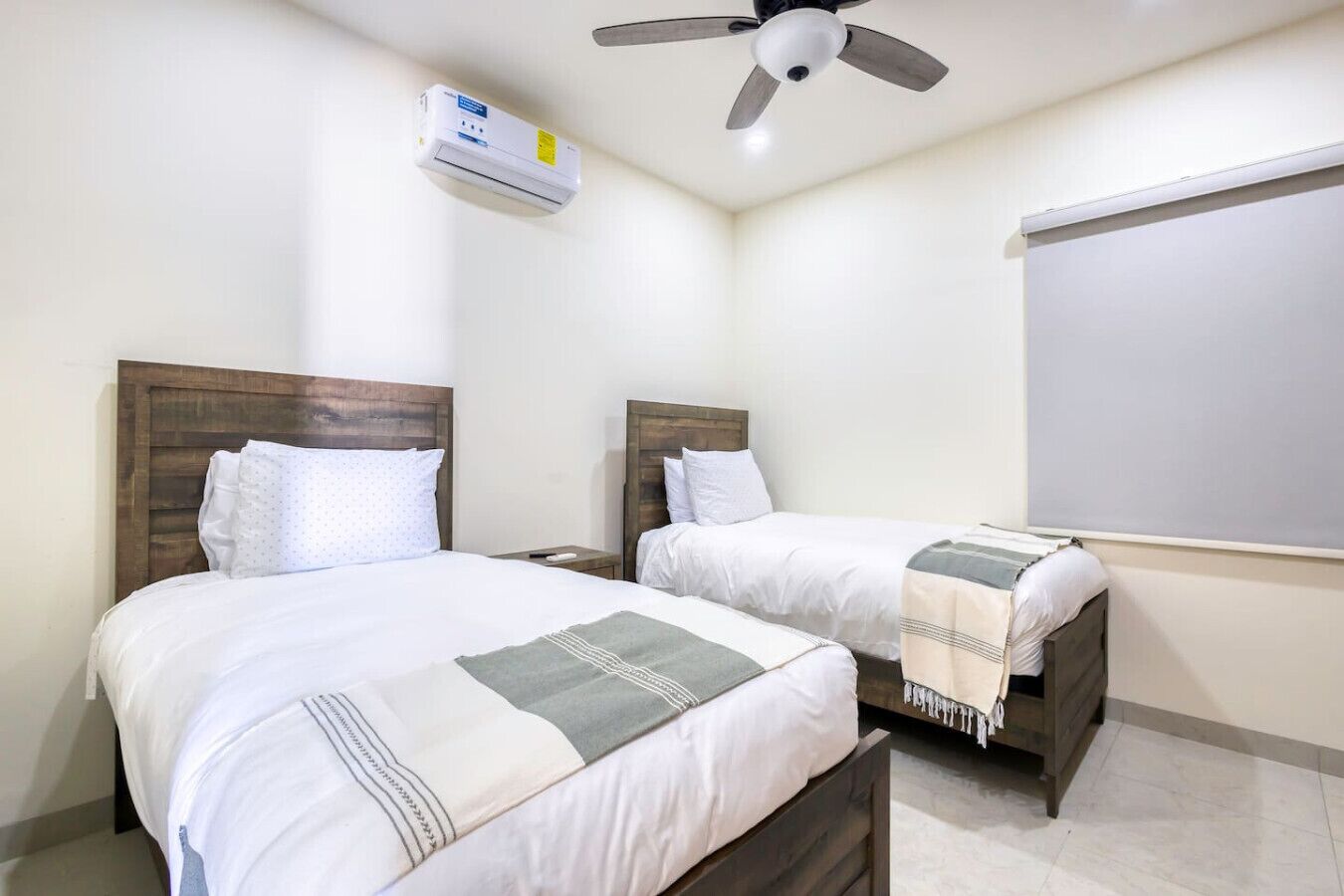 Habitación de hotel con dos camas individuales, ventilador de techo, equipo de aire acondicionado y una ventana con persiana.